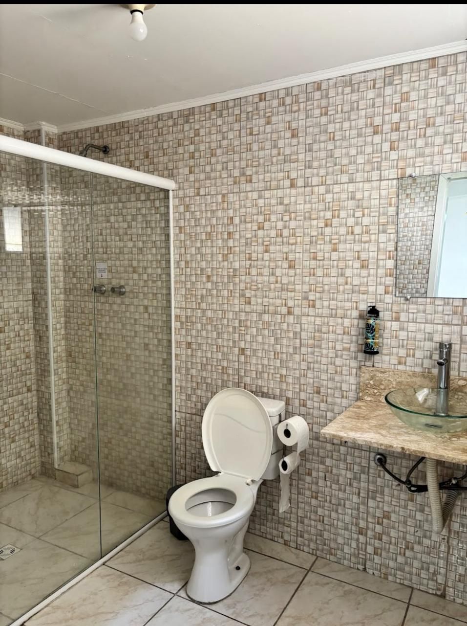 Banheiro com chuveiro, vaso sanitário e pia. Paredes revestidas com azulejos bege