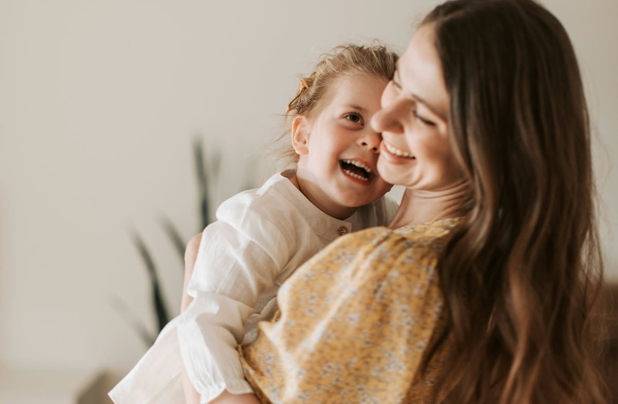 Woman holding and smiling at a laughing child; both have long hair, light background.