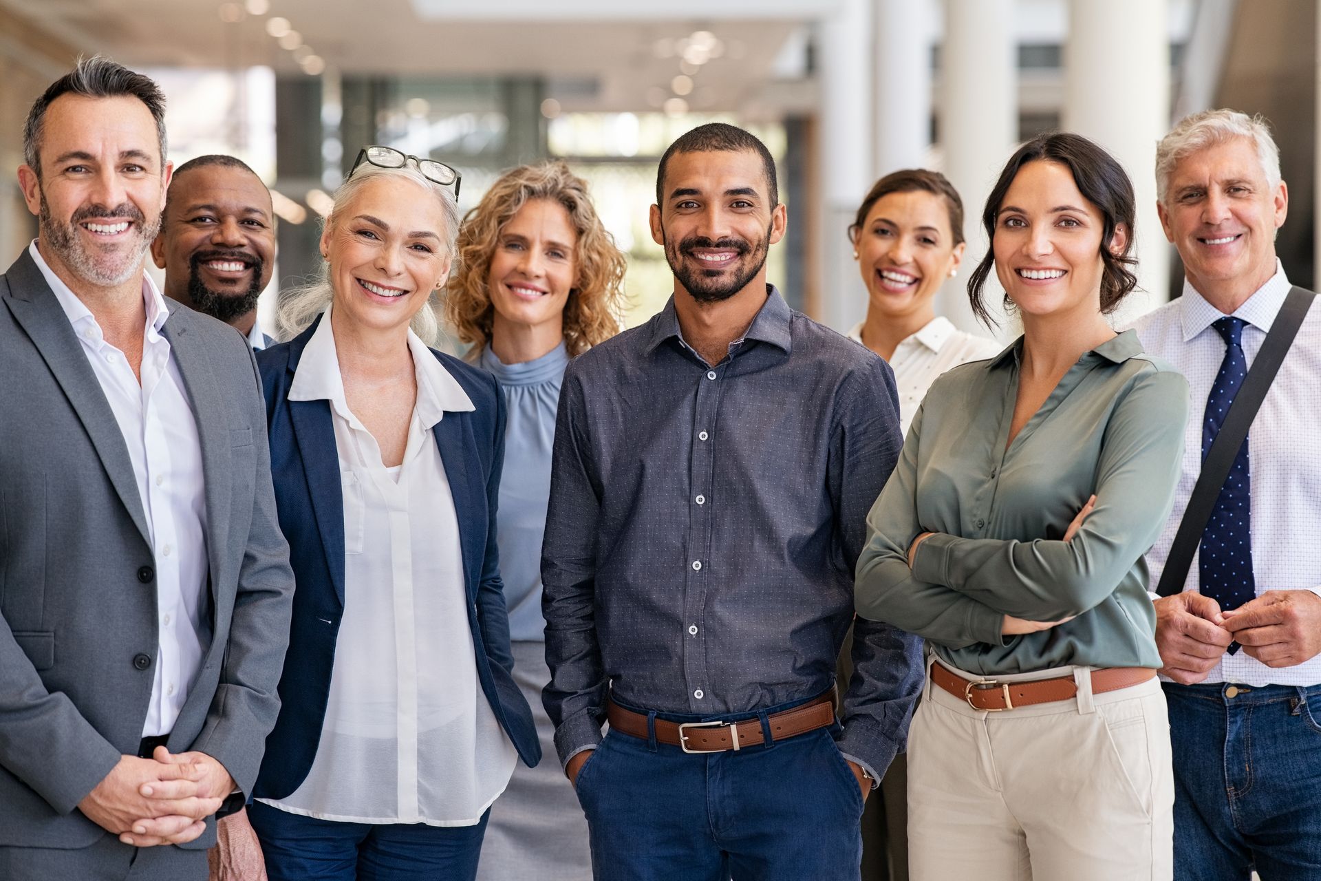 Group of diverse business professionals smiling at the camera, standing in an office hallway.