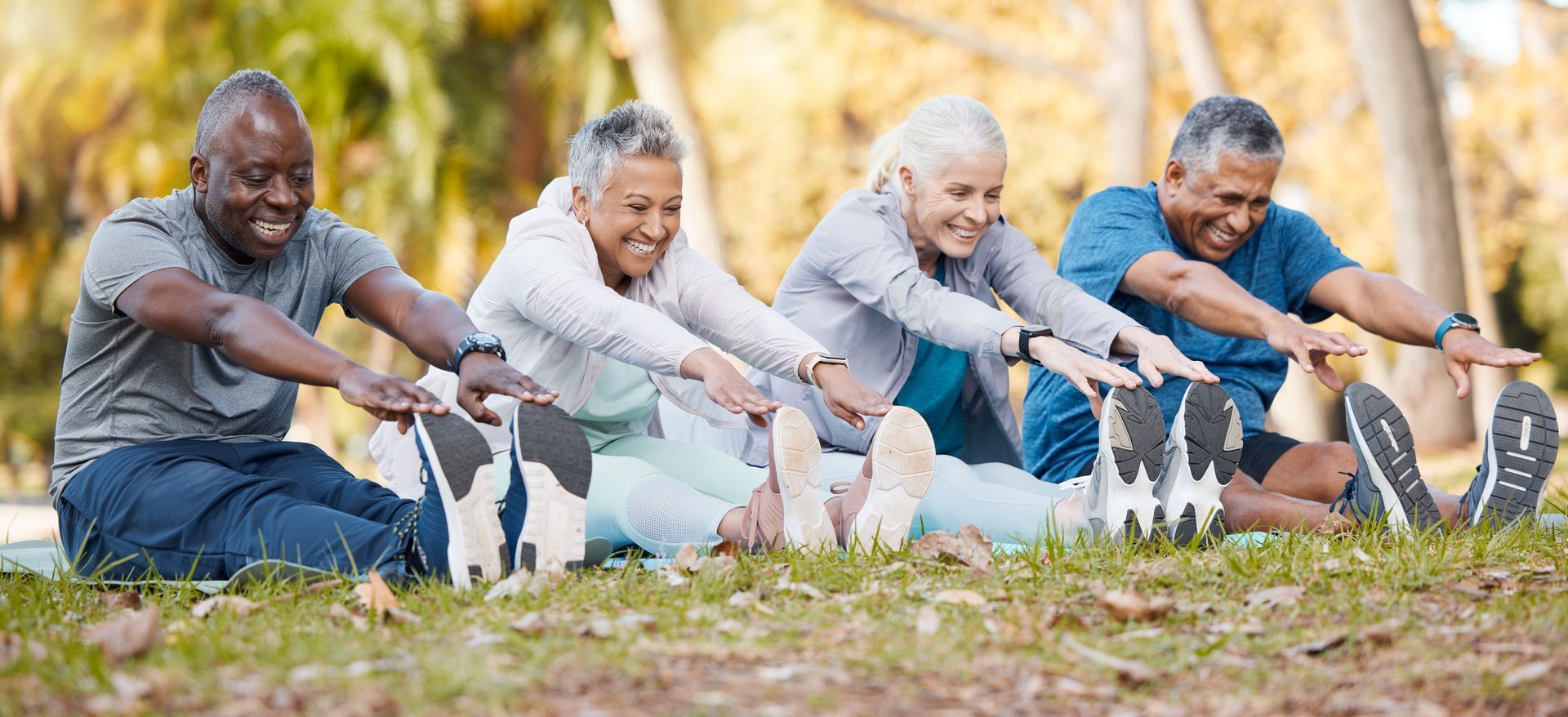 Four older adults stretching their legs, sitting on grass outdoors. They're smiling, reaching for their toes in exercise clothing.