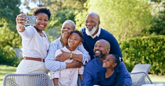 Multi-generational Black family smiling for a selfie outdoors. People hug, and a woman holds the phone.
