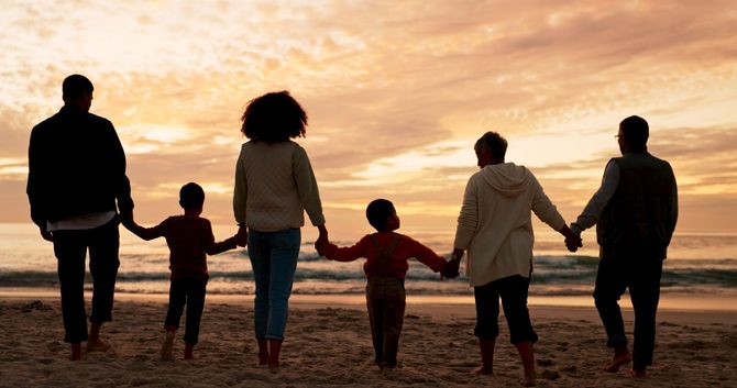 A multigenerational family holds hands on a beach at sunset. Silhouettes of adults and children face the ocean.