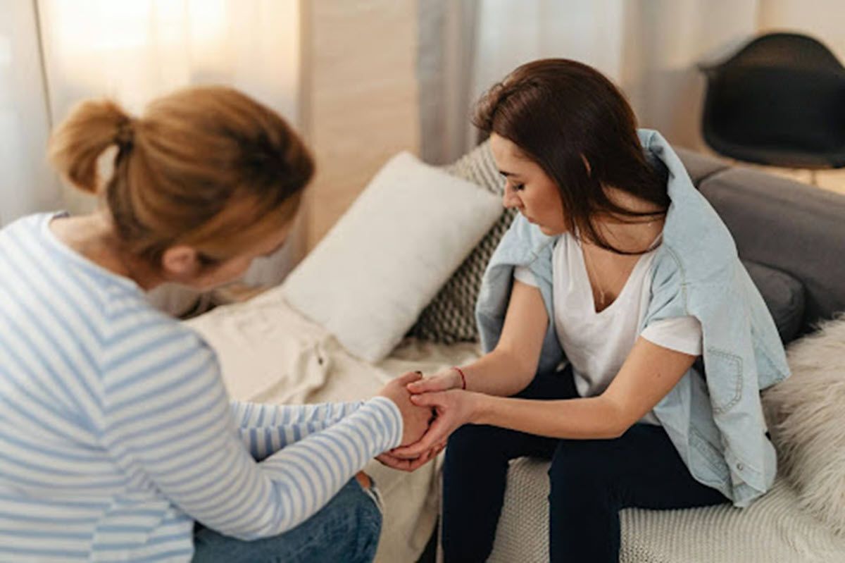 Woman comforting another, holding hands; indoors on a couch.
