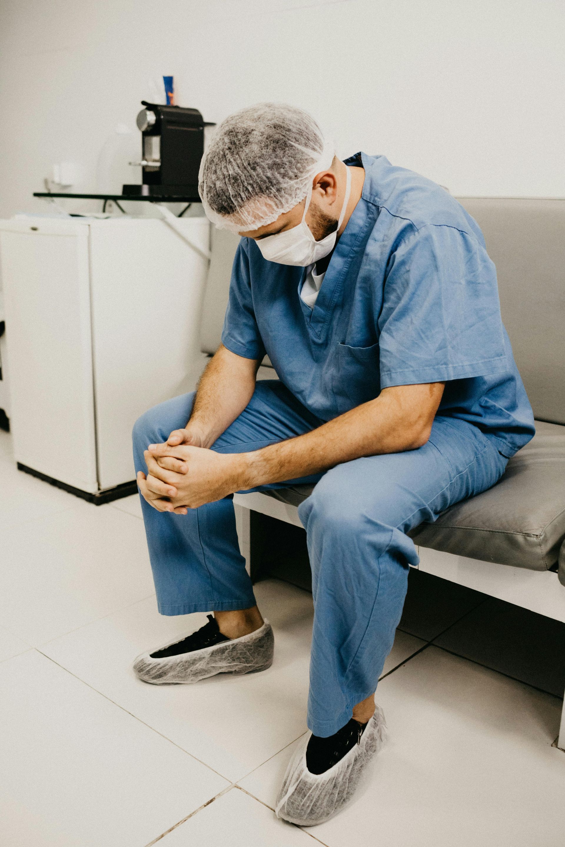 A healthcare worker in blue scrubs, mask, and hairnet sits slumped on a gray couch, head bowed, in a sterile room.