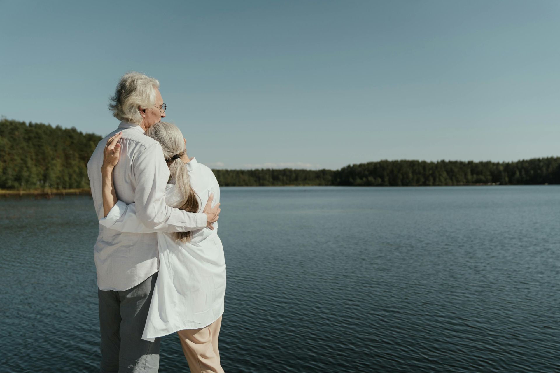 Elderly couple embracing, looking out over a tranquil lake with forest backdrop under a clear blue sky.