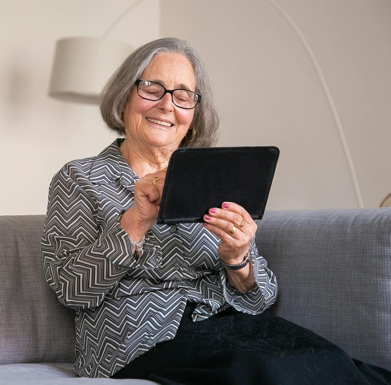 An elderly woman with glasses smiles while using a tablet on a gray couch, indoors.