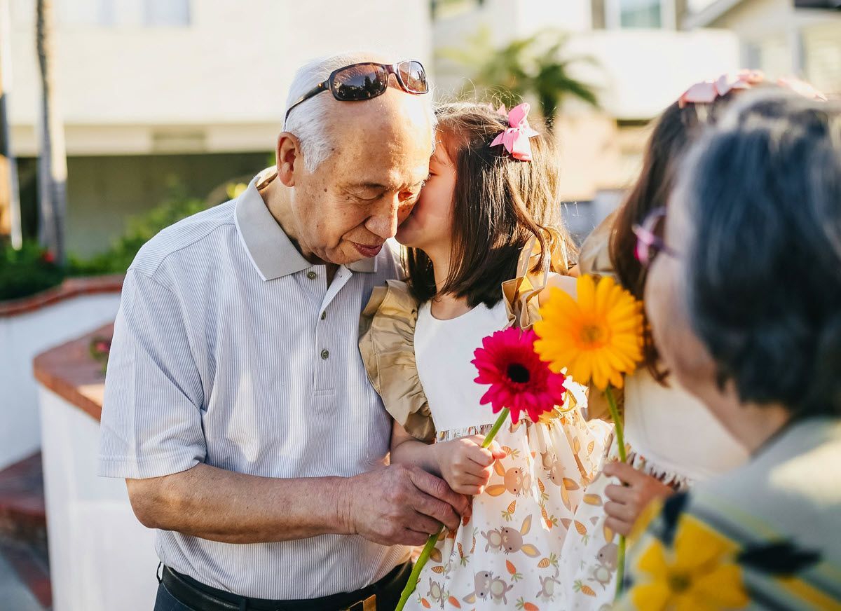 Grandfather gets kissed by a child holding a flower; another person stands nearby, all outdoors.
