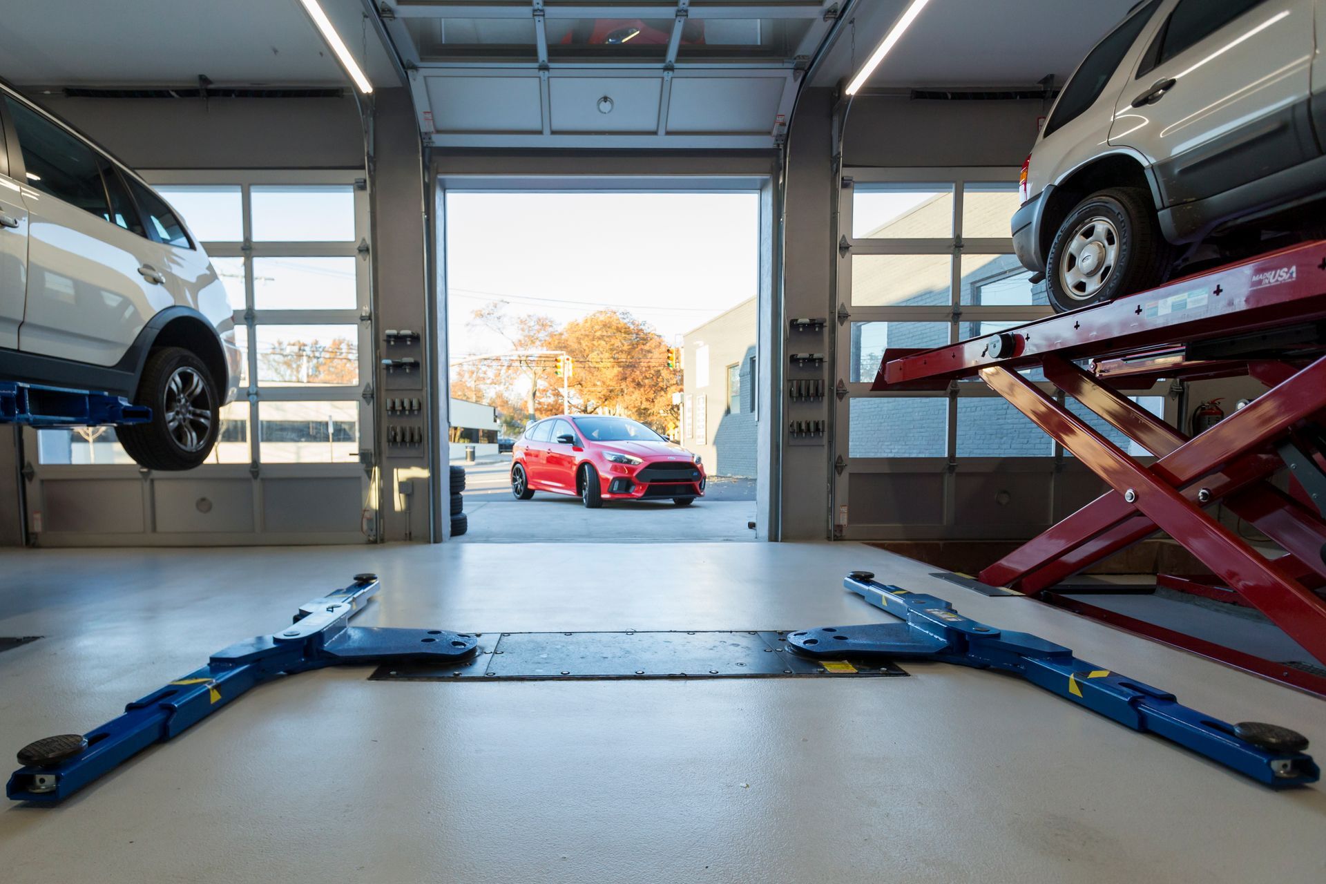 Car repair shop with cars on lifts, open garage door, red car visible outside. | Plandome Service Center
