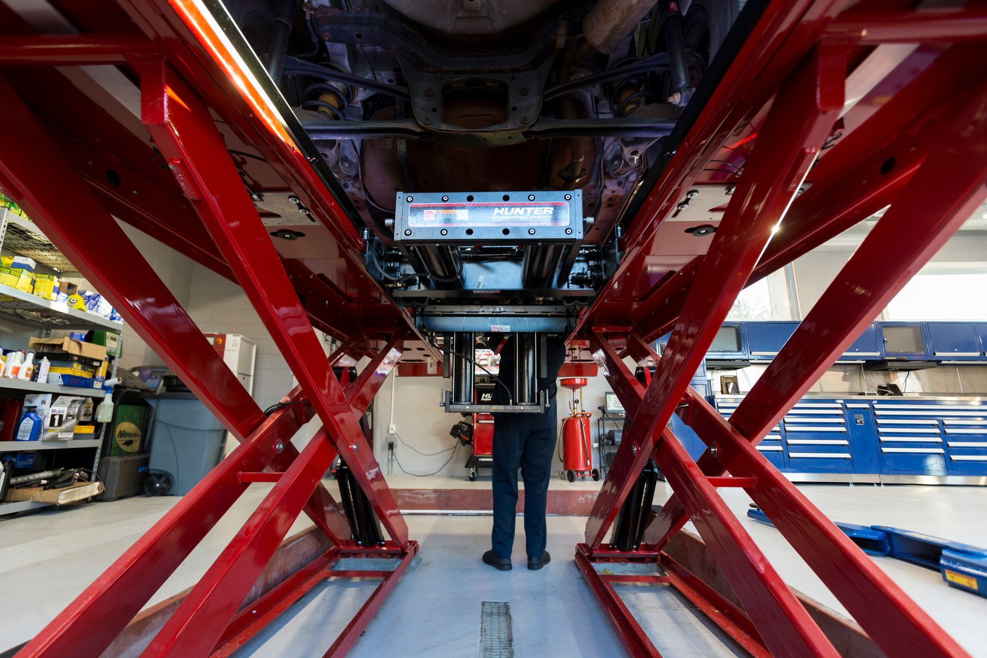 Person under a car on a red lift in a garage; looking at undercarriage. Blue cabinets and tools in background. | Plandome Service Center