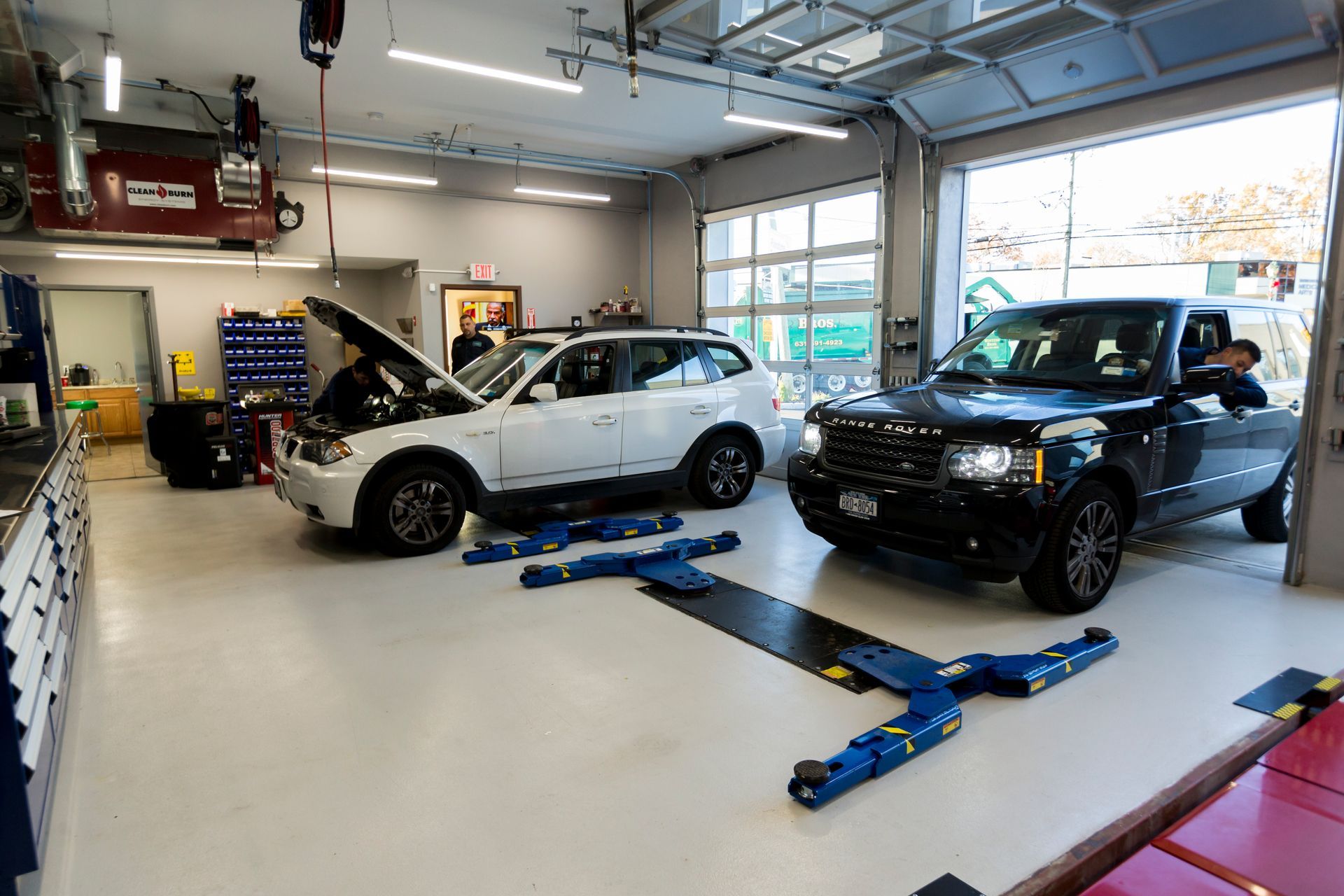 An auto repair shop with a white SUV and black Range Rover. A mechanic works on the white SUV. | Plandome Service Center