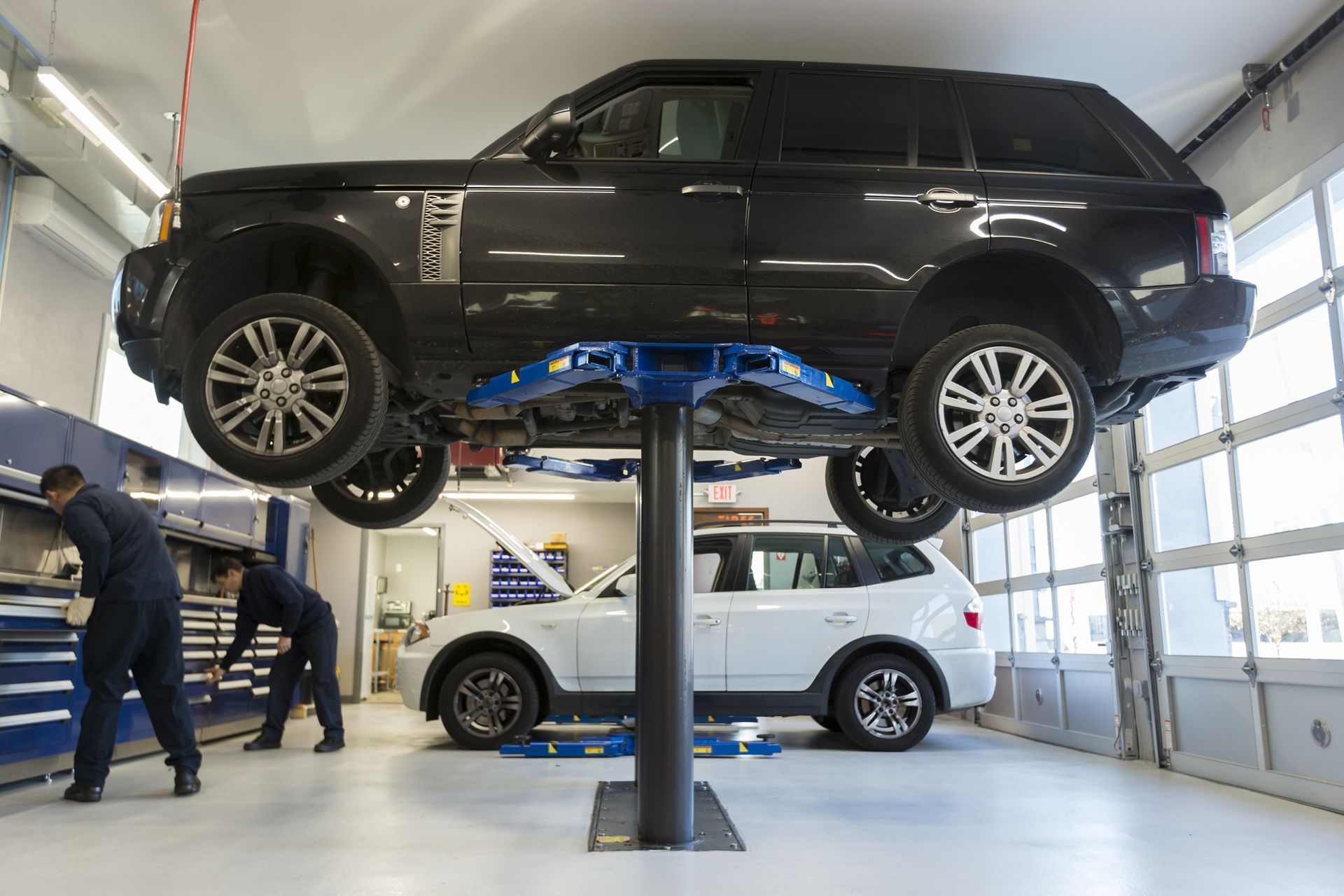 Black SUV on a lift, white car below, mechanics working in a shop. | Plandome Service Center