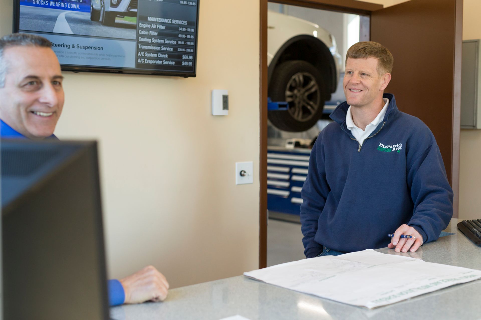 Two men smile, one at a desk, the other standing, in an auto repair shop, car on a lift in the background. | Plandome Service Center