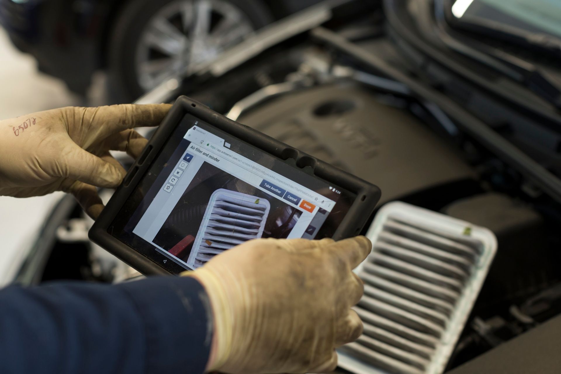 Mechanic holds tablet showing dirty air filter in front of a car engine compartment, hands gloved. | Plandome Service Center