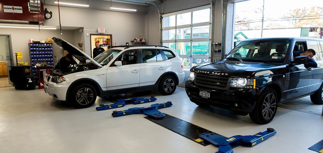 Two cars in a garage, a white SUV with open hood and a black SUV, are on lifts, with a person in each car. | Plandome Service Center
