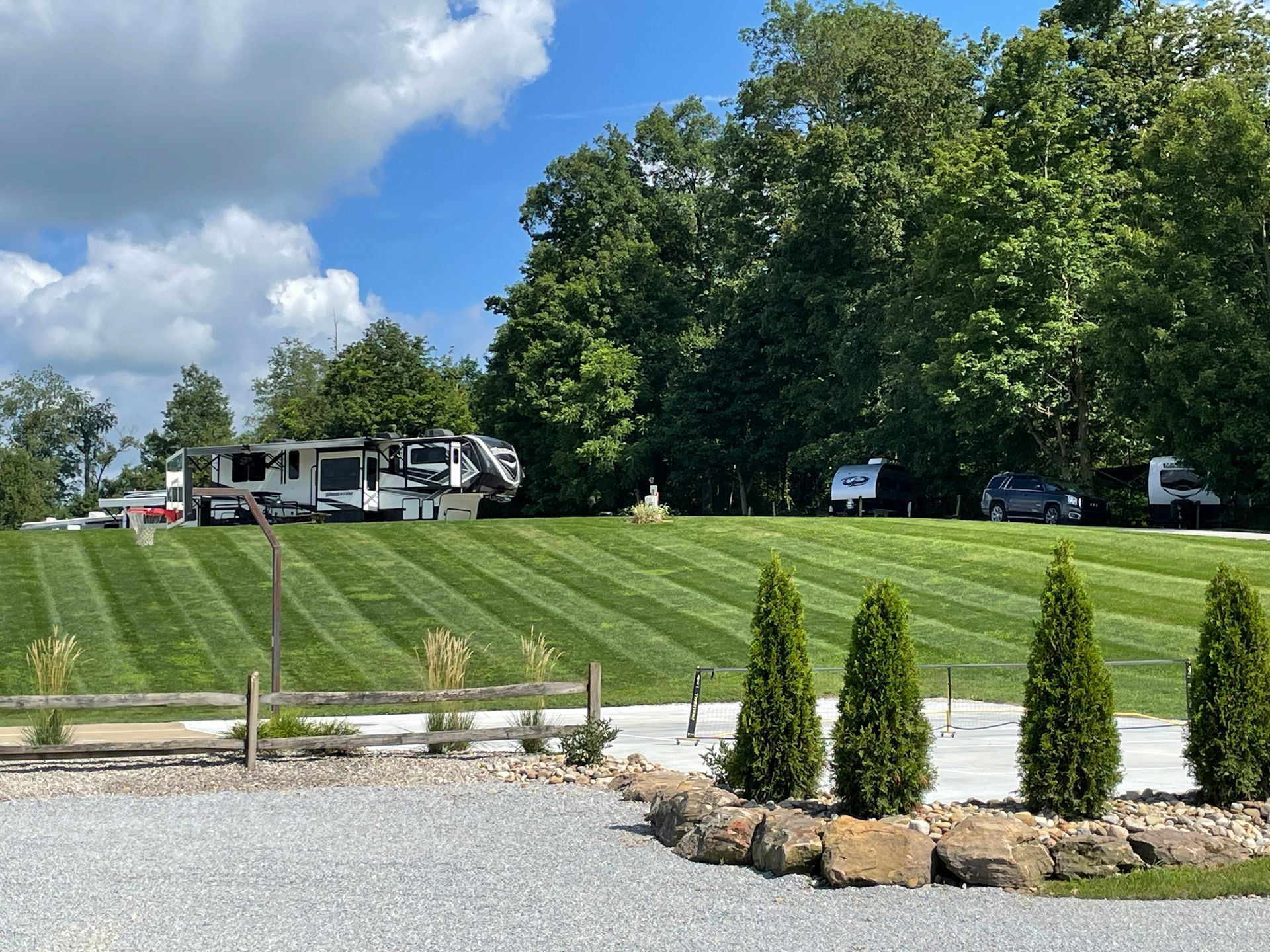 A small building with a gravel garden, wagon decor, and rows of green trees; a lake in the background.