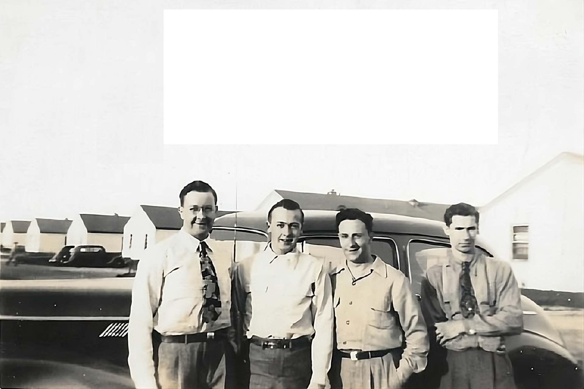 Four men pose in front of a car and houses, likely 1940s. They are wearing collared shirts, ties and slacks. | Ramsey's Service Center