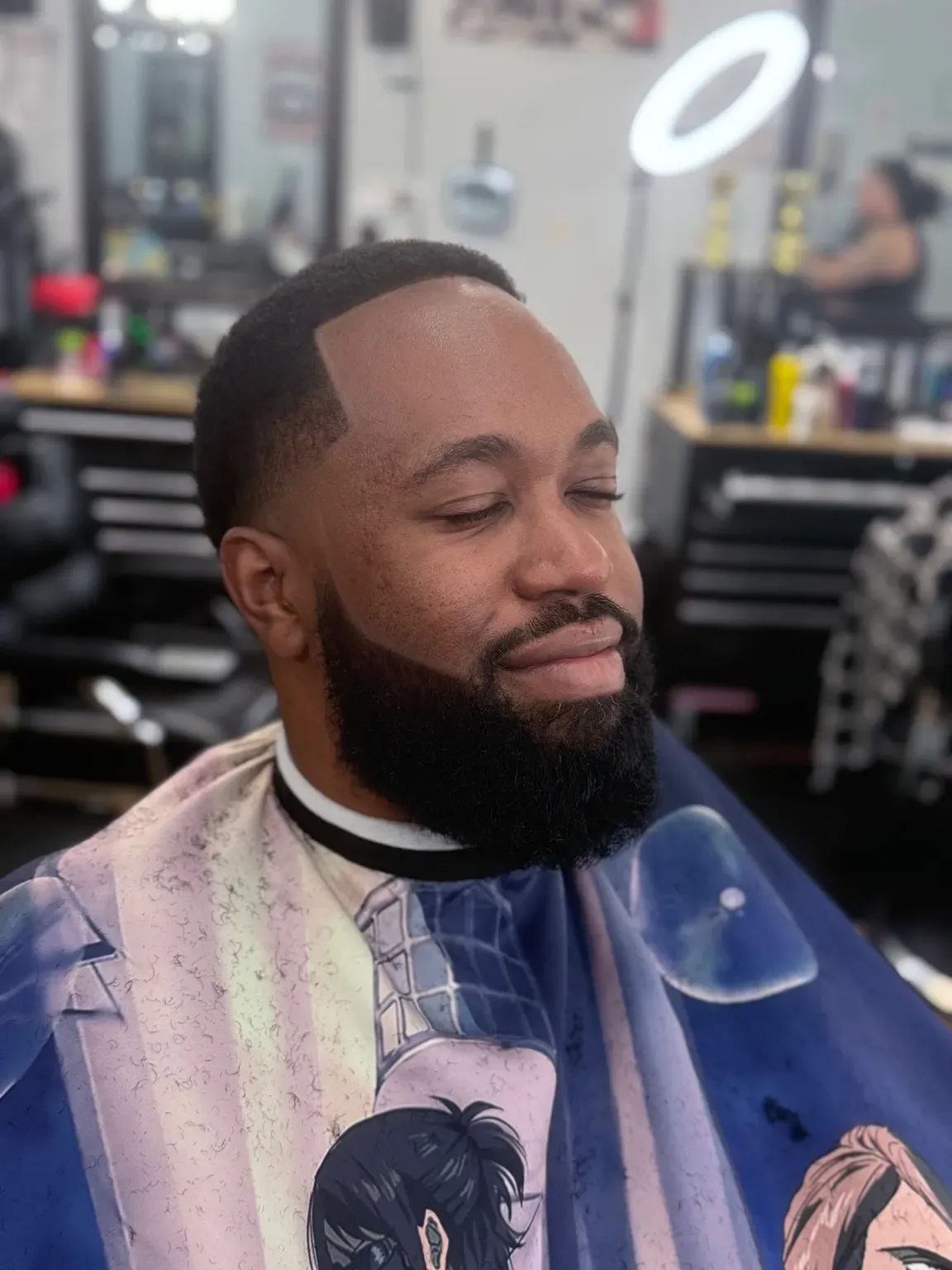 Man with a faded haircut and beard at a barbershop, smiling with closed eyes, wearing a barber cape.