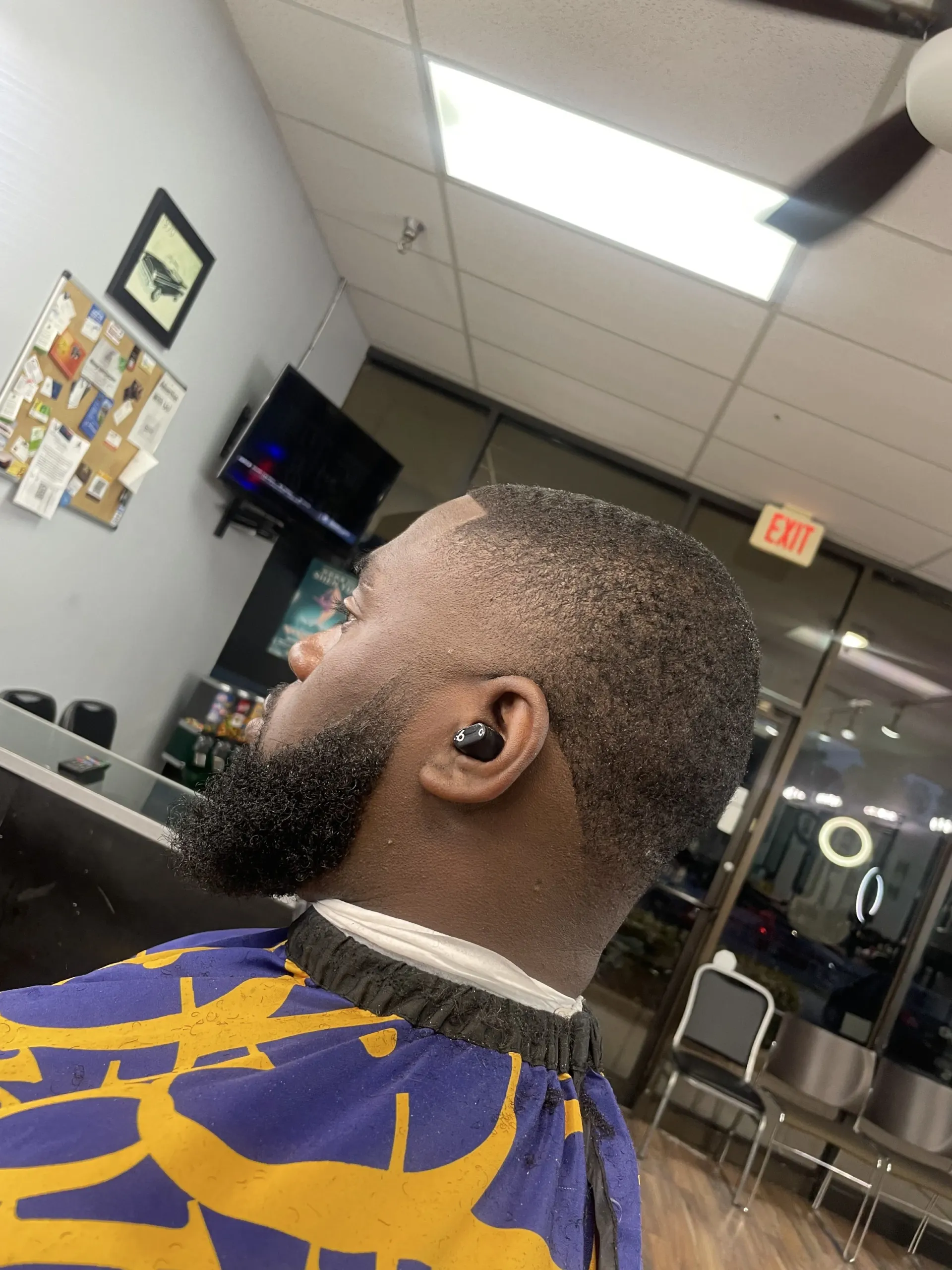 Man with a short haircut and beard wearing a barber cape; inside a barber shop with a TV and lights.