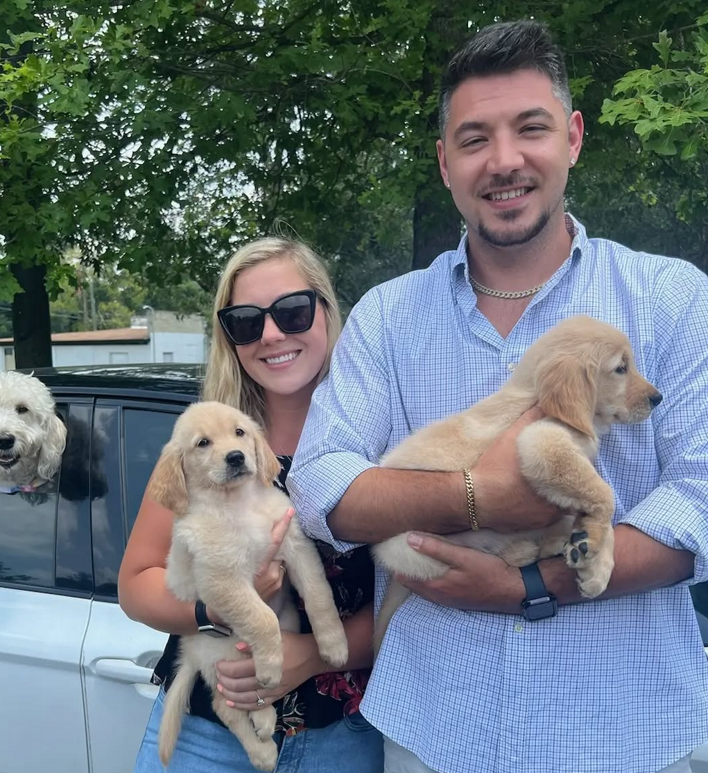 A man and a woman are holding two puppies in front of a car.