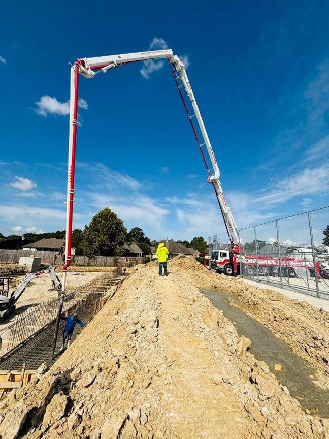 Concrete being poured on a construction site. A person in a yellow vest stands on a dirt mound, under the extended boom. Blue sky. Concrete being poured on a construction site. A person in a yellow vest stands on a dirt mound, under the extended boom. Blue sky.