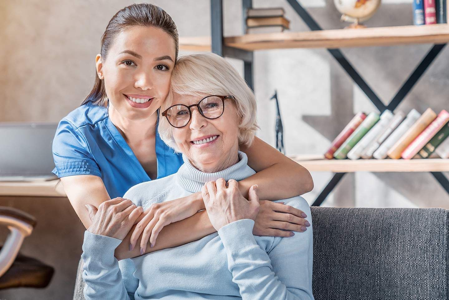 Woman in blue uniform hugs smiling older woman indoors.