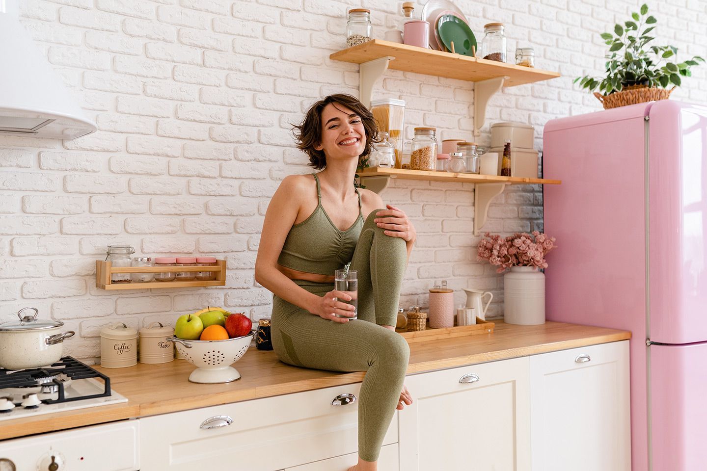 Woman in olive green workout clothes smiles, sitting on a kitchen counter, holding water bottle.