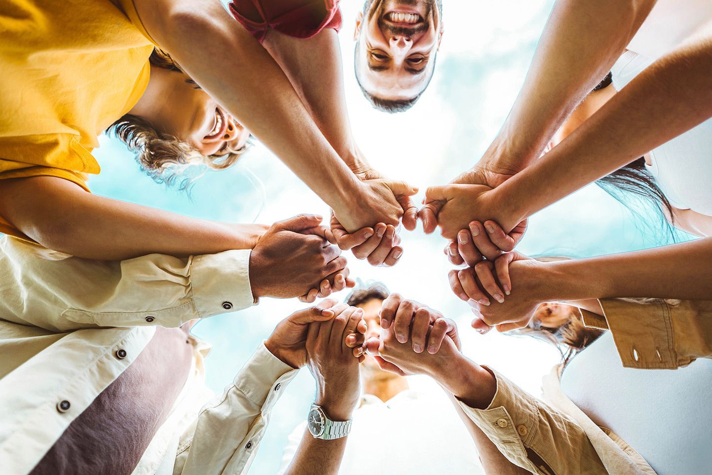 Hands clasped together in a circle, viewed from below, against a bright sky, symbolizing unity and teamwork.