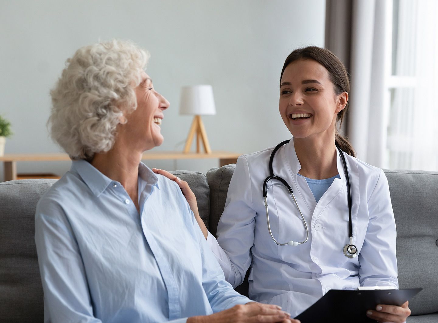 Doctor smiles at and puts her hand on an elderly patient's shoulder, as they sit together indoors.