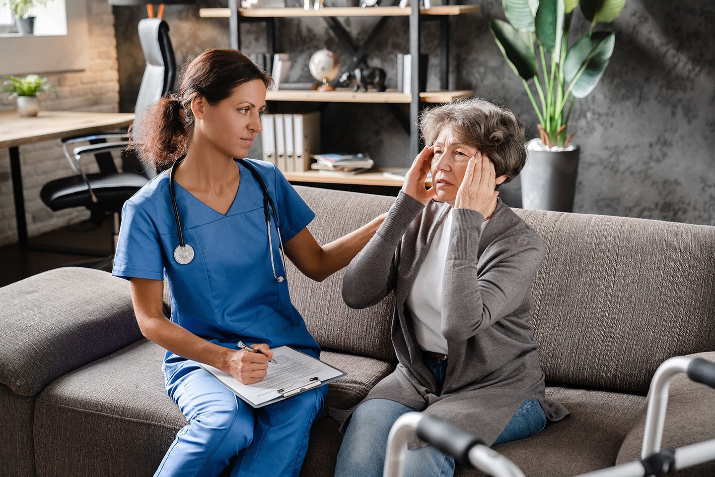 Nurse in blue scrubs comforts an older person on a couch, holding her head.