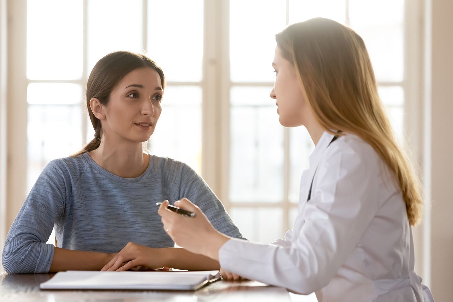 Woman in blue top consults with a doctor in a white coat. They are sitting at a table with papers, indoors.