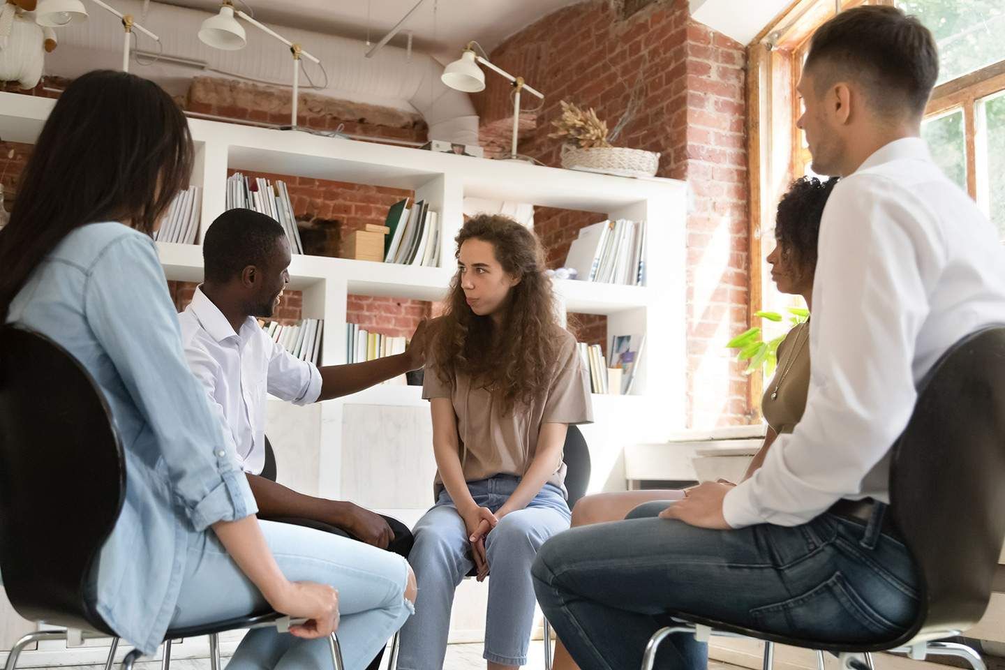Group therapy session: People seated in a circle, offering support.  Setting: Bright room with bookshelves and a window.