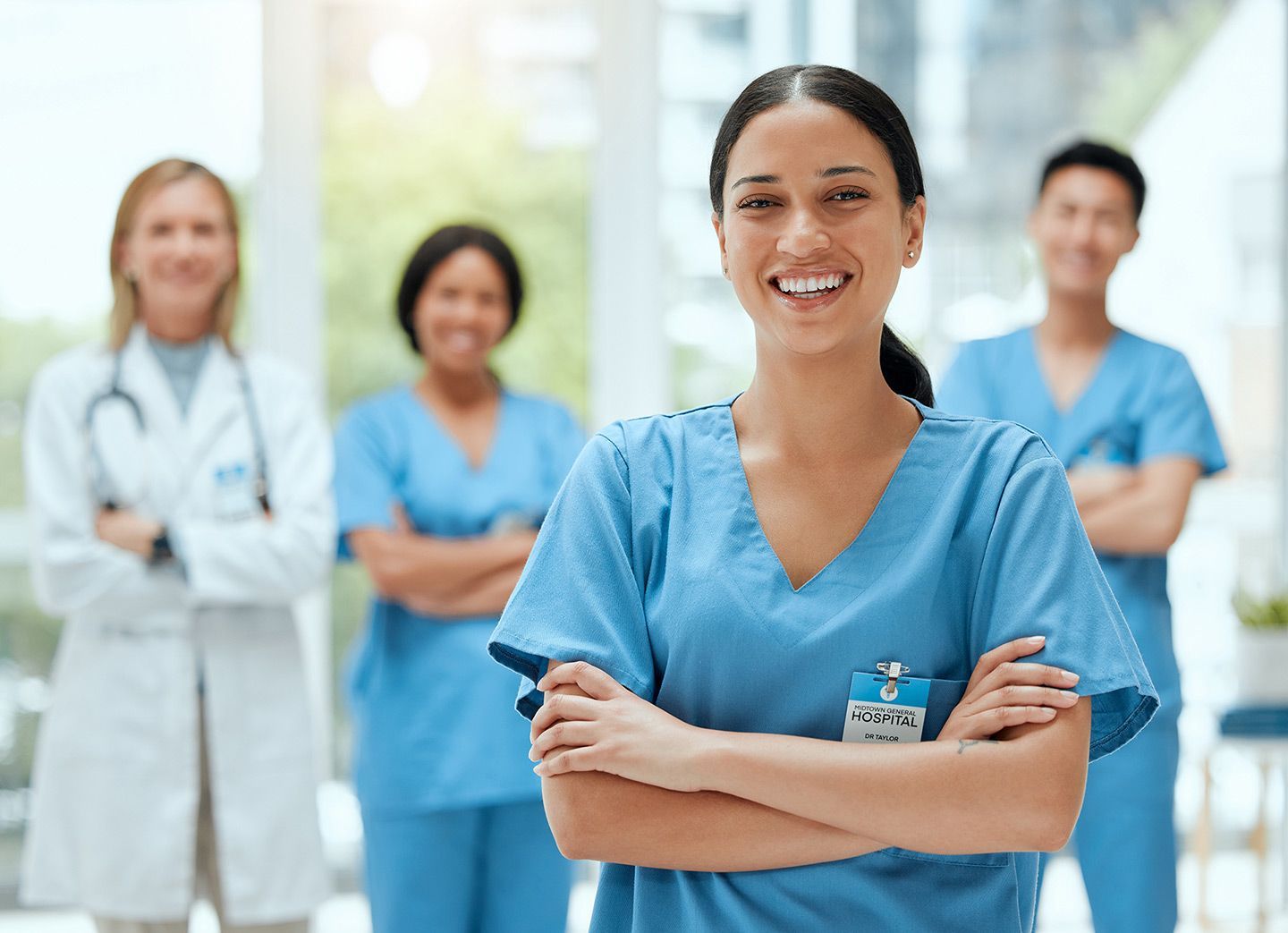 Medical team smiling, arms crossed. Woman in blue scrubs in the foreground. Others in scrubs and lab coat in background.