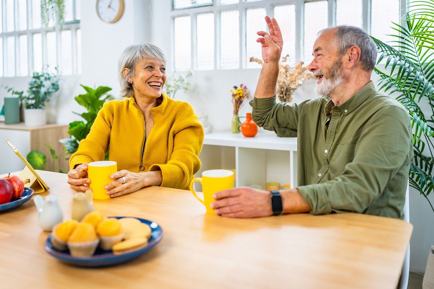 Couple at table, smiling, drinking from yellow mugs. Man raising hand, woman wearing yellow jacket.