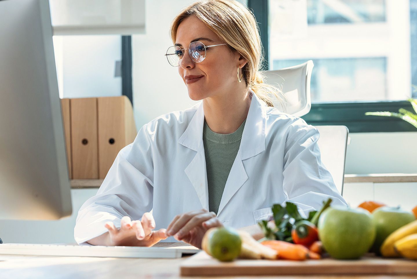 Woman in lab coat looks at computer, fruit and vegetables on table.
