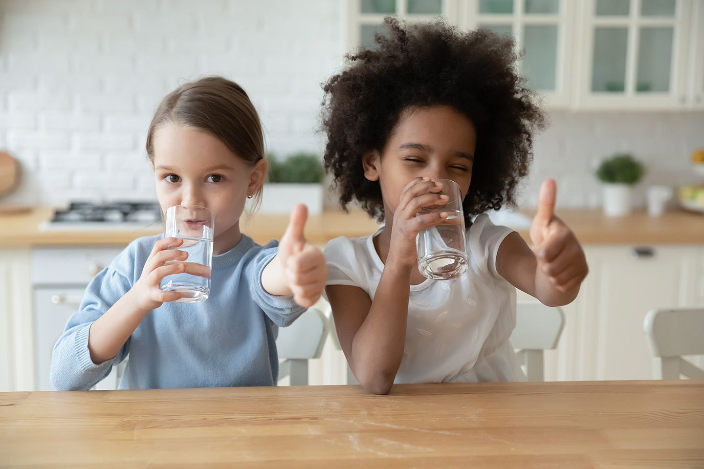 Two children drinking water and giving thumbs up in a kitchen.