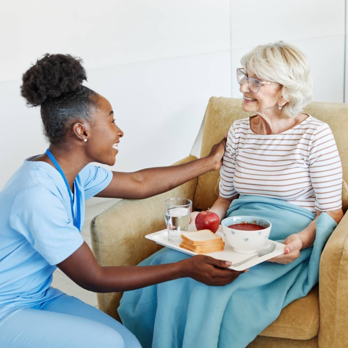 Caregiver offering a tray of food to an older adult seated in a chair.