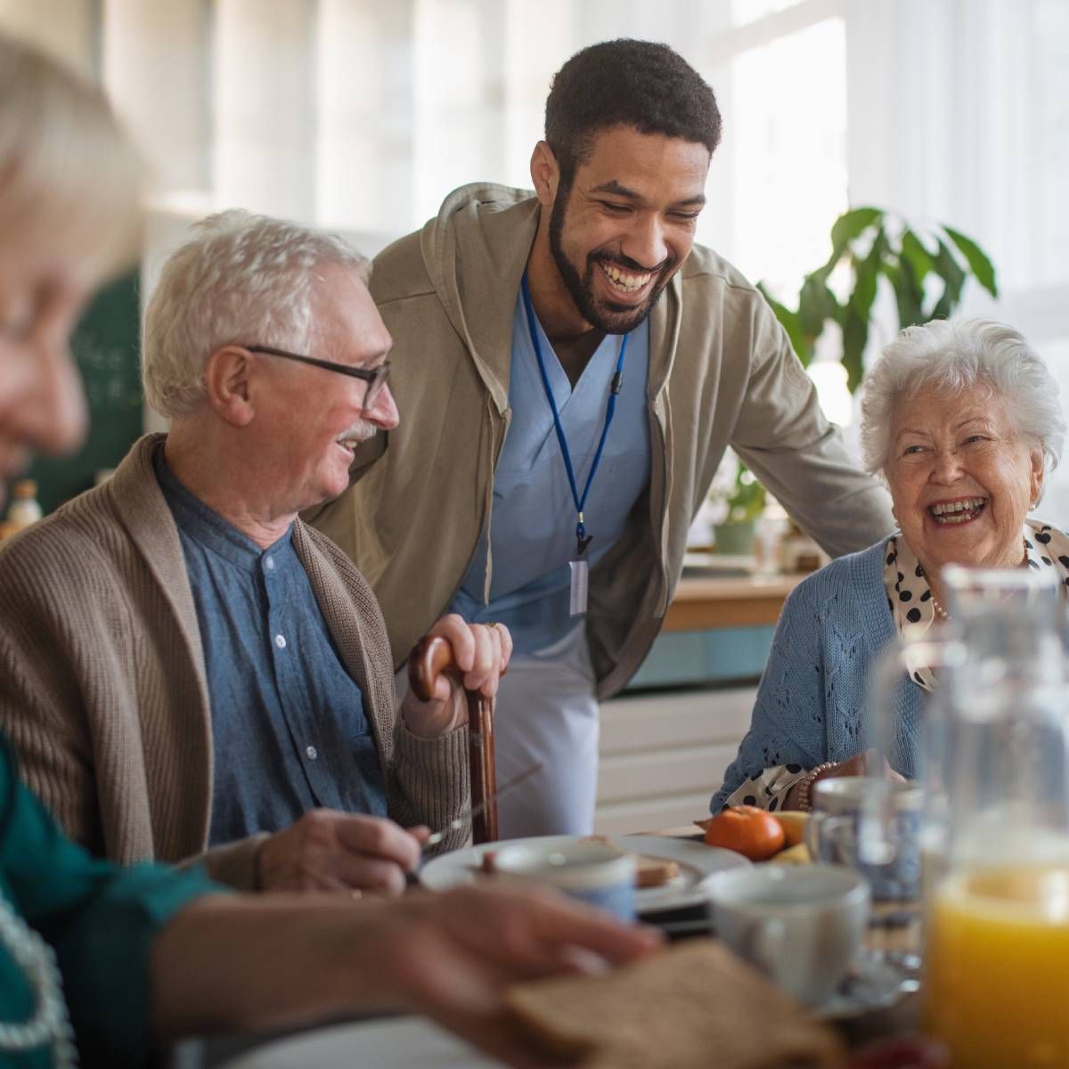 Caregiver smiles at seniors at a breakfast table, some laughing.