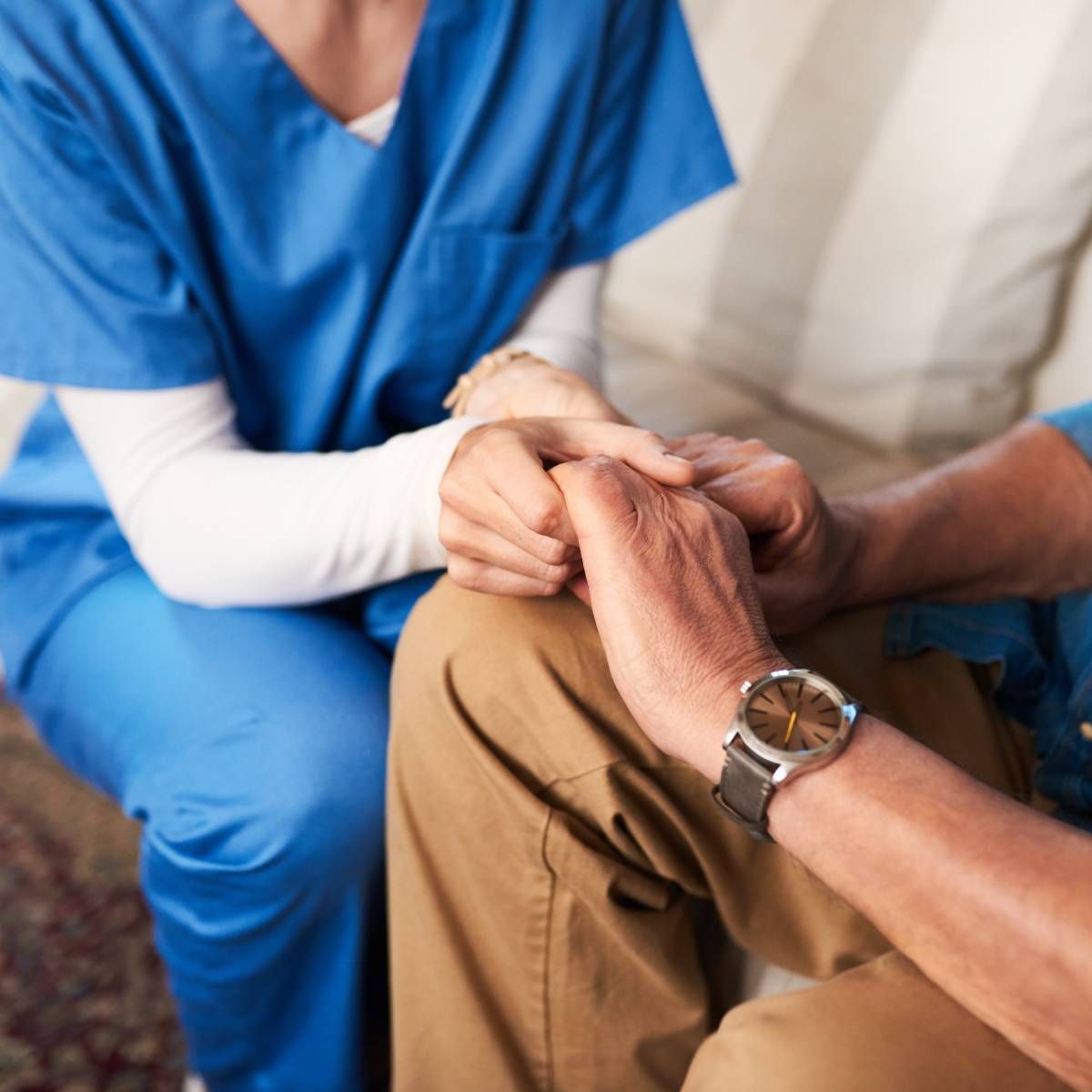 A person in blue scrubs holds the hands of another person wearing a watch.