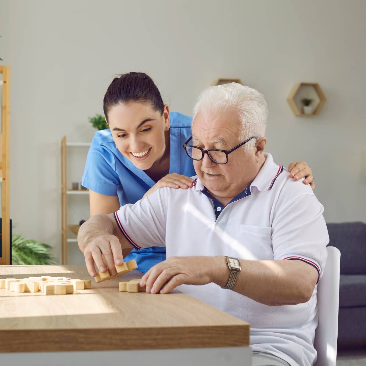Caregiver helps an older person play a wooden block game at a table. Smiling woman wears scrubs, man wears glasses.
