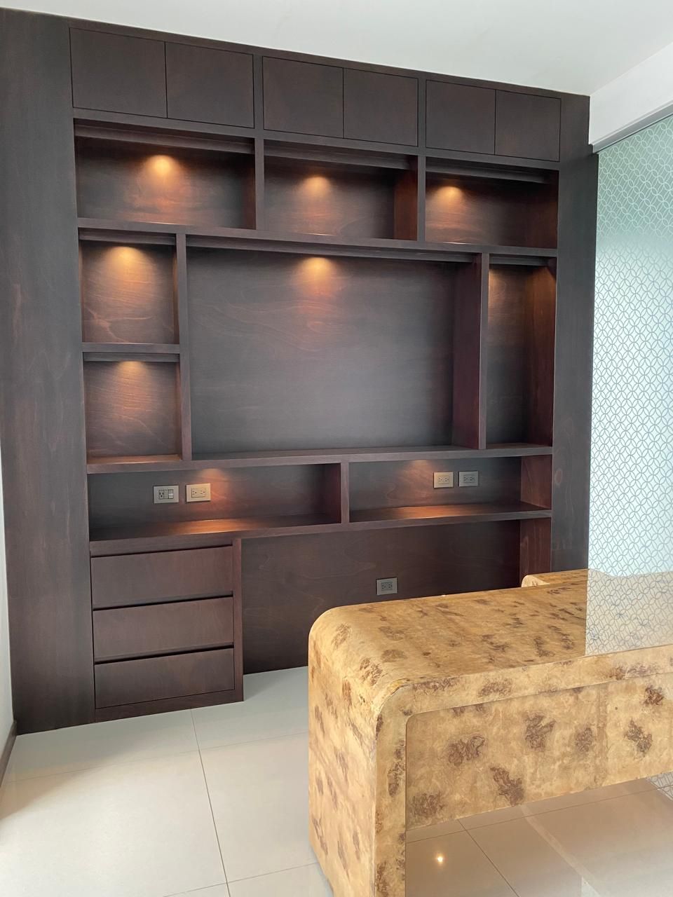 Estantería empotrada de madera oscura con cajones y luces, encima de una zona de estar con otomana de color tostado.