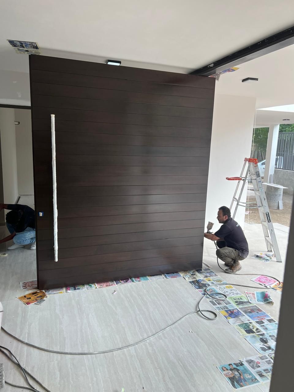 Persona rociando pintura sobre una gran puerta de madera oscura dentro de un edificio en construcción.