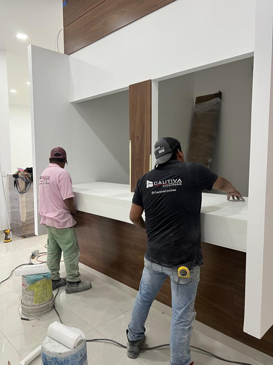 Dos trabajadores instalando paneles de madera en una pared blanca; obra de construcción interior.