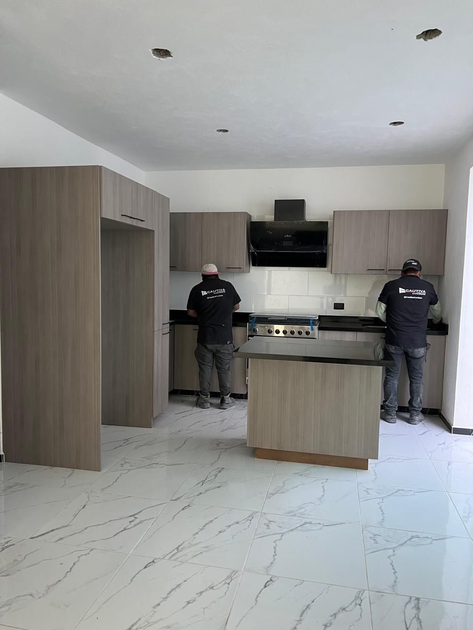 Dos trabajadores instalando gabinetes de cocina en una cocina moderna con gabinetes de madera clara y pisos de mármol.