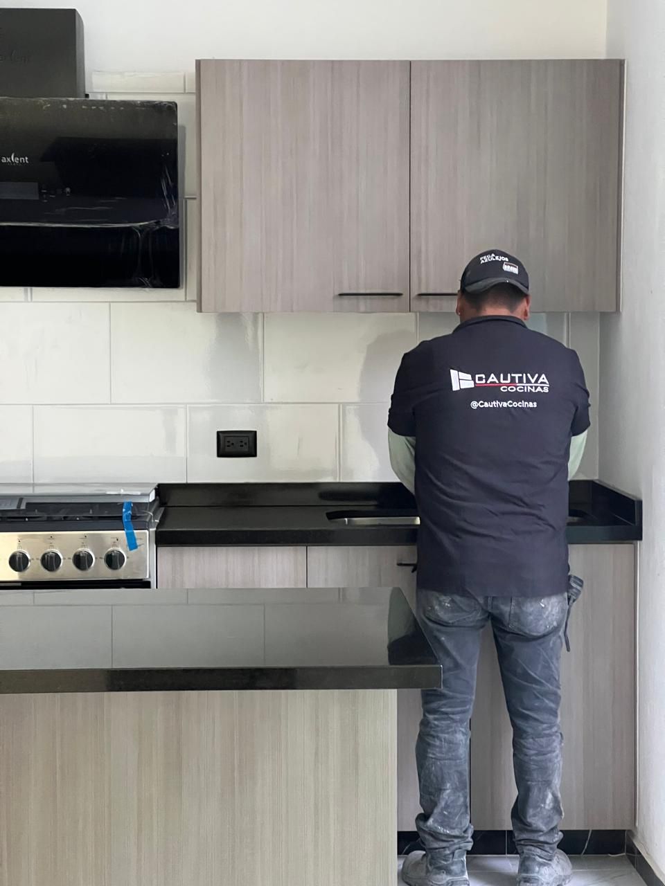 Hombre instalando una encimera de cocina. Gabinetes grises, encimera negra, estufa y campana extractora.