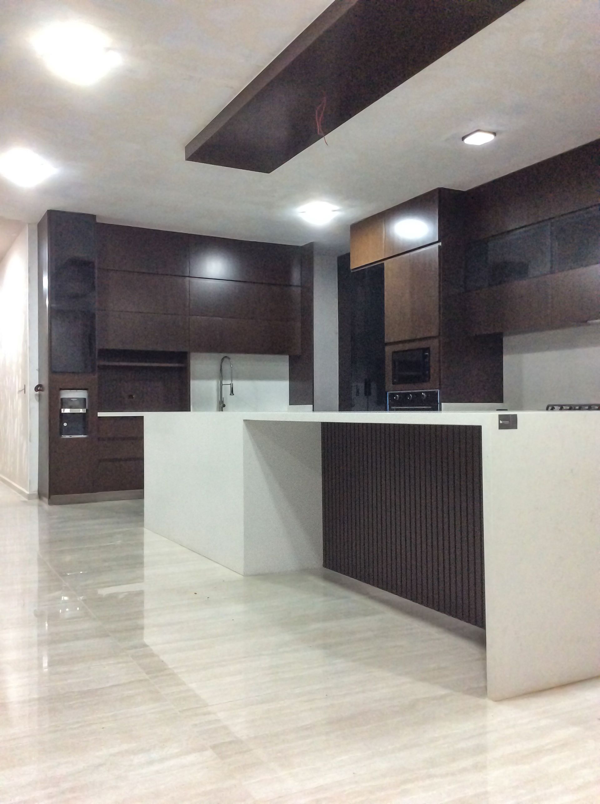 Cocina moderna con muebles de madera oscura, encimeras blancas e isla. Iluminación cenital brillante y suelos de color claro.