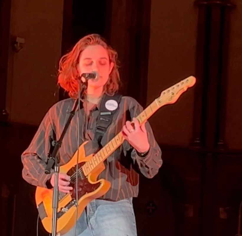 Person playing electric guitar on stage, singing into a microphone. Dark shirt, jeans, warm lighting.