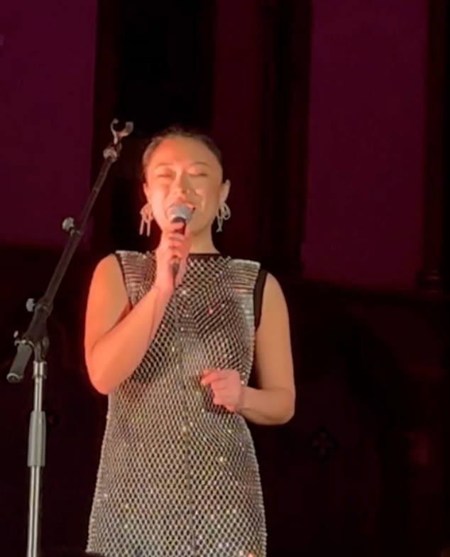 Woman singing into a microphone onstage, wearing a sparkly silver dress.