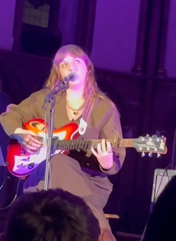 Woman singing and playing electric guitar on stage with purple lighting.