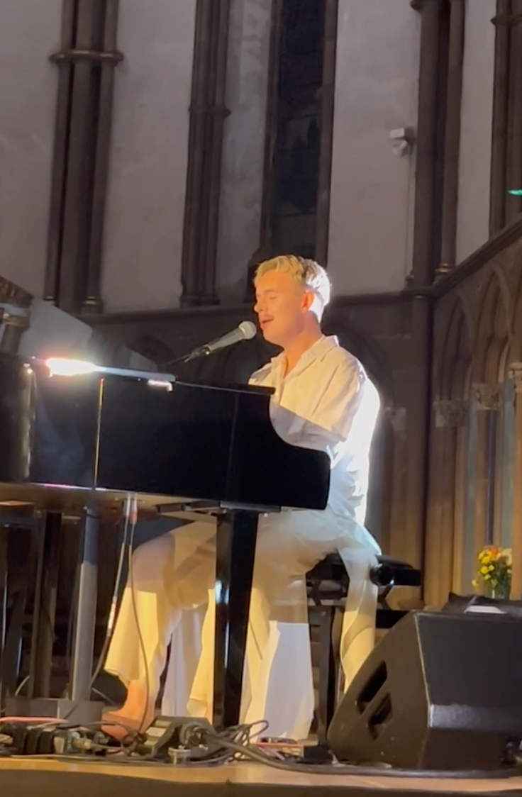 A person singing and playing piano on a stage, lit by a spotlight. Bare feet. White attire.