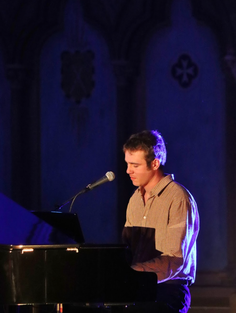Man playing piano, singing into a microphone in a blue-lit church. He wears a beige shirt and sits at a grand piano.